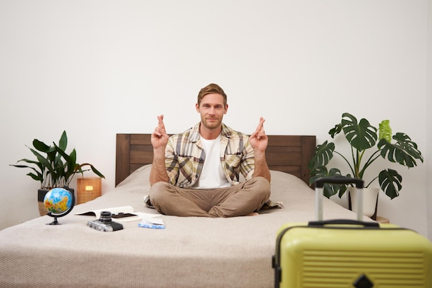 Photo gratuite portrait d'un jeune homme méditant avec les doigts croisés priant faisant un vœu emballant une valise pour partir