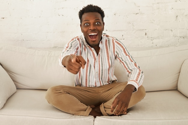 Portrait de jeune homme à la peau sombre choqué émotionnel se détendre à la maison, assis confortablement sur le canapé, ayant excité le regard ravi, pointant le doigt avant à la caméra