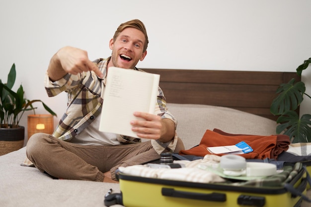 Photo gratuite portrait d'un jeune homme pointant vers sa liste d'articles emballant des vêtements en vacances montrant un cahier