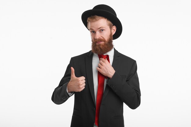 Portrait d'un jeune homme rousse élégant avec une longue barbe taillée posant dans des vêtements élégants à la mode, montrant les pouces vers le haut en signe d'approbation, va acheter ce costume et ce chapeau