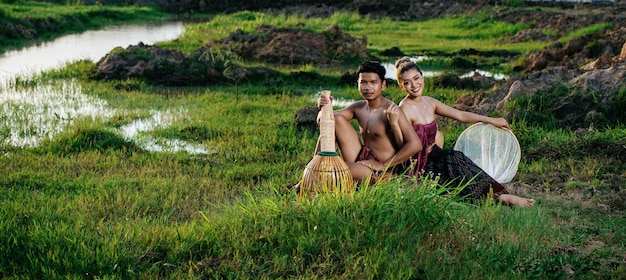 Photo gratuite portrait jeune homme seins nus portant des pagnes dans un mode de vie rural assis près d'une belle femme avec un piège à pêche en bambou