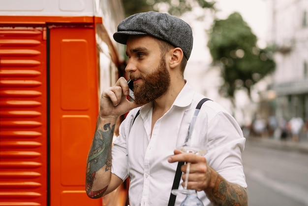 Portrait de jeune homme tatoué barbu en gros plan appelle au téléphone dans la rue. Un mec romantique en chemise blanche, casquette et bretelles. Peaky Blinders. démodé, rétro.