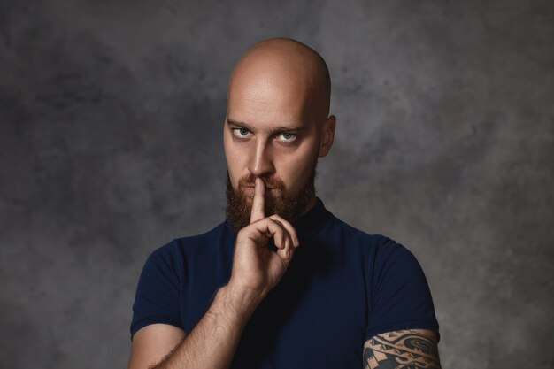 Portrait de jeune homme tatoué sérieux avec la tête rasée et la barbe touffue avec une expression faciale menaçante stricte, tenant le doigt sur ses lèvres, disant de garder la bouche fermée