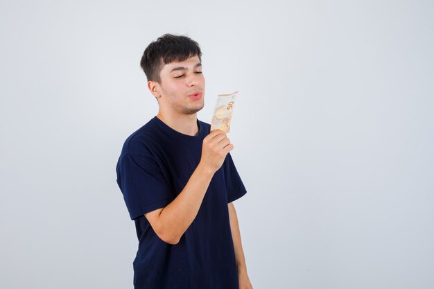 Portrait de jeune homme tenant des billets en euros, souffle de l'air baiser en t-shirt noir et à la vue de face confiante