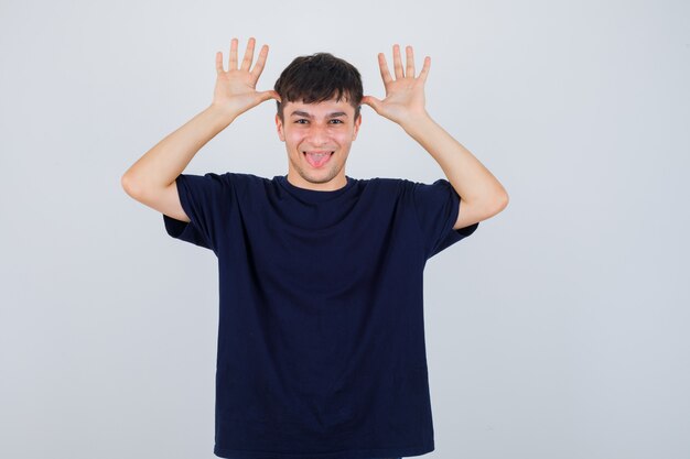 Portrait de jeune homme tenant la main près de la tête comme les oreilles, qui sort la langue en t-shirt noir et à la vue de face amusée