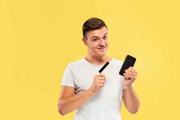 Portrait de jeune homme tenant smartphone et carte de crédit isolé sur mur jaune