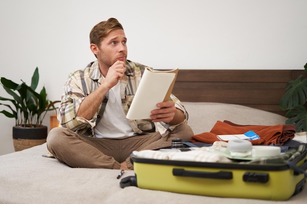 Portrait d'un jeune homme touriste réfléchi planifiant ses vacances assis avec une valise ouverte sur le lit
