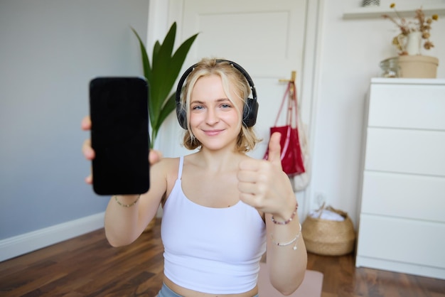 Photo gratuite portrait d'un jeune instructeur de fitness dans des écouteurs montrant l'écran d'un smartphone et les pouces en l'air