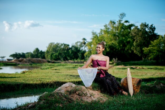 Portrait Jeune jolie femme asiatique dans de beaux vêtements traditionnels thaïlandais au champ de riz, elle s'asseyant près de l'équipement de pêche