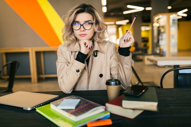 Portrait de jeune jolie femme ayant une idée, assis à table en trench-coat travaillant sur ordinateur portable