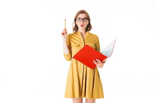 Portrait de jeune jolie femme à lunettes et robe jaune debout avec dossier rouge et crayon dans les mains tout en regardant étonnamment à huis clos sur fond blanc isolé