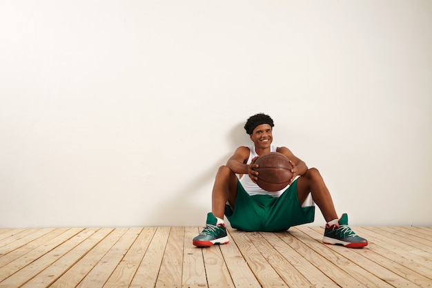 Un portrait d'un jeune joueur souriant assis sur le plancher en bois contre un mur blanc tenant un vieux ballon de basket brun