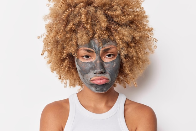 Portrait d'une jeune mannequin aux cheveux bouclés bouleversée applique un masque d'argile sur le visage pour éliminer les pores subit des procédures de beauté quotidiennes porte un t-shirt isolé sur fond blanc. Concept de soins du visage