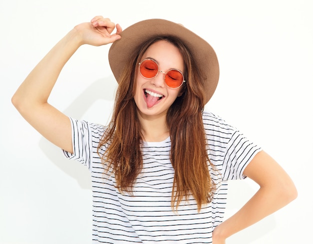 Portrait de jeune mannequin femme élégante qui rit dans des vêtements d'été décontractés en chapeau brun avec maquillage naturel isolé sur mur blanc. Montrant sa langue