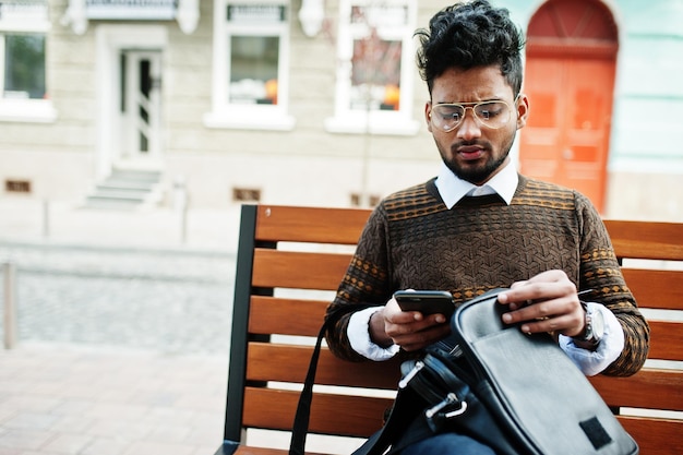 Photo gratuite portrait de jeune mannequin indien élégant pose dans la rue assis sur un banc avec sac à main et smartphone