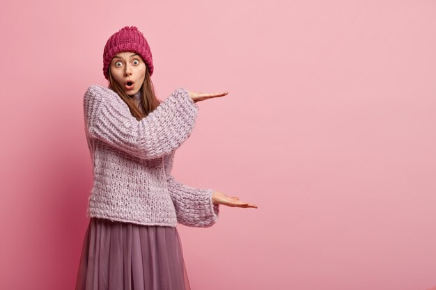 Portrait de jeune modèle féminin surpris forme un gros objet avec les deux mains, impressionné par la taille, habillé en chapeau, pull et jupe longue, isolé sur un mur rose. Concept de personnes et de mesure