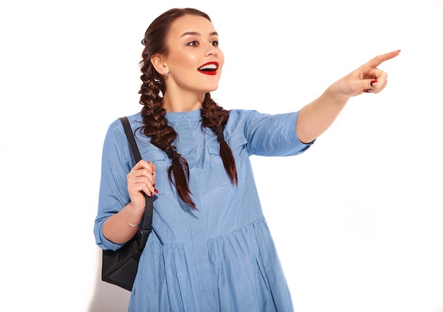 Portrait de jeune modèle femme souriante heureuse avec maquillage lumineux et lèvres rouges avec deux nattes en mains en été robe bleue colorée et sac à dos isolé. Pointant vers le côté