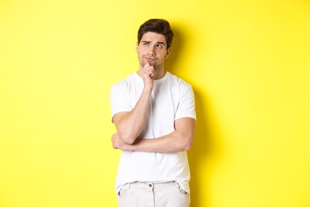 Portrait de jeune modèle masculin pensant, regardant le coin supérieur gauche et faisant un choix, debout près de l'espace de copie, fond jaune.