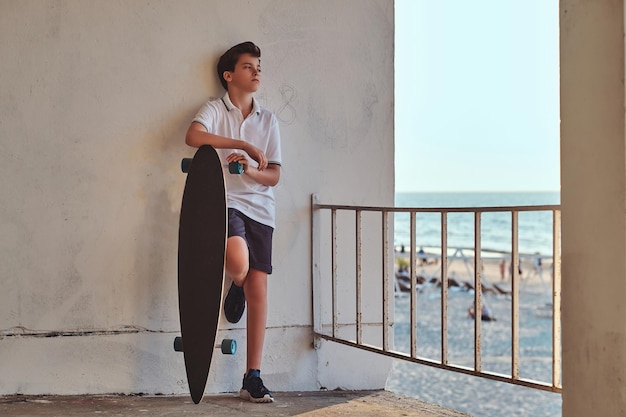 Portrait d'un jeune patineur vêtu d'un t-shirt et d'un short appuyé sur un mur et tenant une planche à roulettes sur fond de côte.