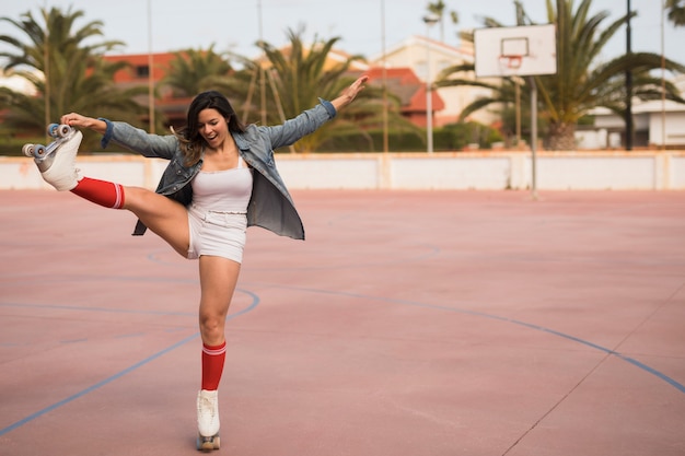 Photo gratuite portrait d'une jeune patineuse souriante en équilibre sur une jambe, debout sur un terrain de basket