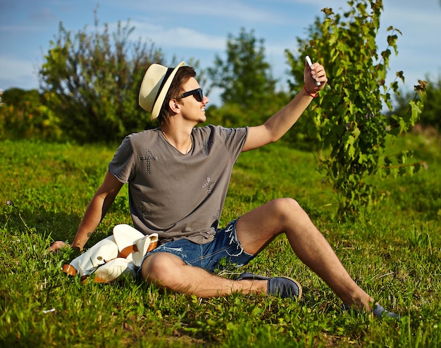 portrait, de, jeune, séduisant, moderne, élégant, homme, dans, désinvolte, tissu, dans, chapeau, dans, lunettes, séance, dans parc, dans, herbe verte, confection, selfie