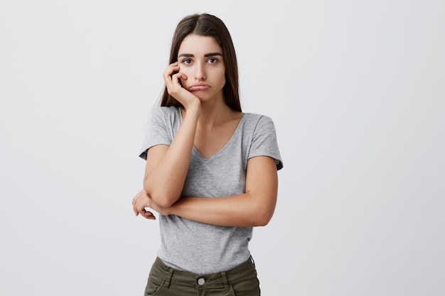 Portrait de jeune séduisante triste charmante étudiante caucasienne aux cheveux longs noirs en tenue grise élégante tenant la tête avec la main avec une expression malheureuse après avoir reçu une mauvaise note