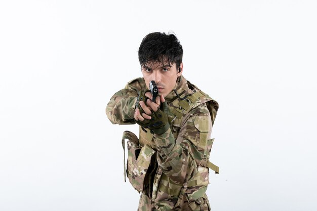 Portrait de jeune soldat en tenue de camouflage avec pistolet sur mur blanc