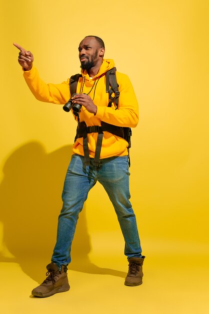 Portrait d'un jeune touriste joyeux avec sac et jumelles isolé sur mur jaune studio