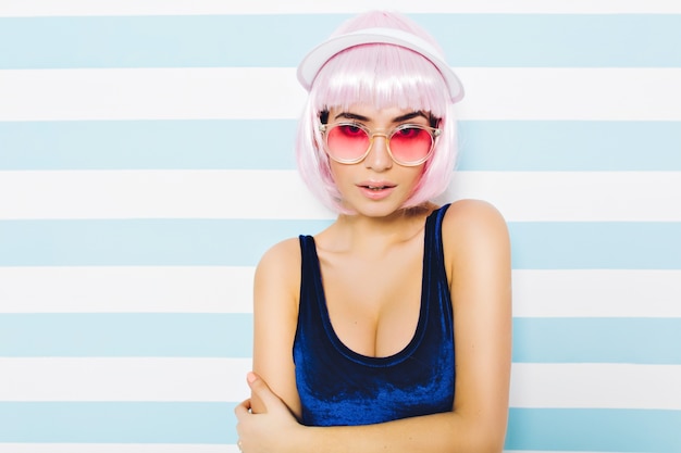 Photo gratuite portrait joli modèle attrayant d'été en maillot de bain bleu avec coupe de cheveux rose, lunettes de soleil roses à la recherche sur le mur blanc bleu rayé. jeune femme sexy, bonne humeur, fête à la plage.
