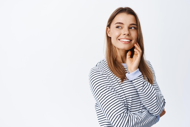 Portrait d'un joli modèle féminin regardant de côté l'espace de copie du logo de l'entreprise touchant le visage avec un maquillage de lumière naturelle et souriant debout heureux sur fond blanc