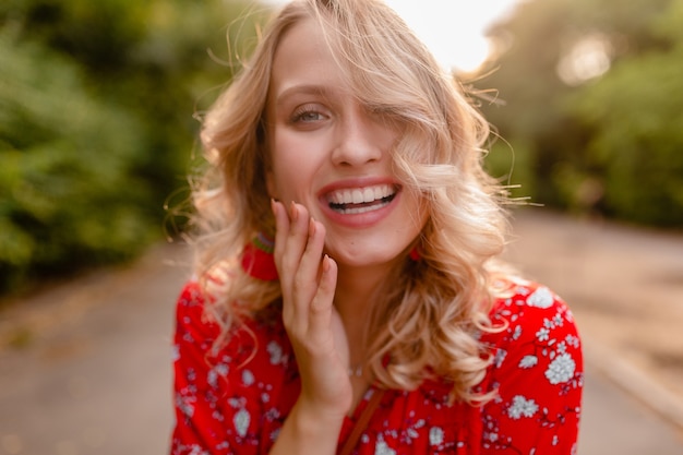 Portrait de jolie blonde élégante femme souriante en chemisier rouge tenue de mode d'été dans le parc style boho portant des boucles d'oreilles souriant