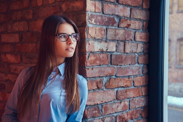 Portrait d'une jolie brune charmante à lunettes et chemise bleue appuyée contre un mur de briques dans une pièce au design loft.