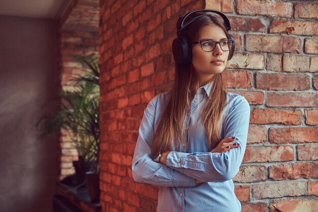 Portrait d'une jolie brune charmante en lunettes et chemise bleue bras croisés et écoutant de la musique sur un casque appuyé contre un mur de briques dans une pièce au design loft.