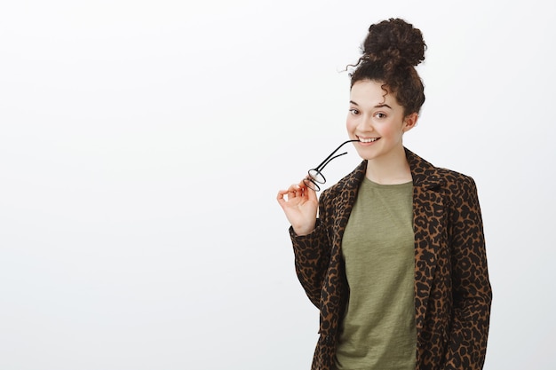 Portrait de jolie étudiante brune aux cheveux bouclés, mordant le bord de lunettes et souriant