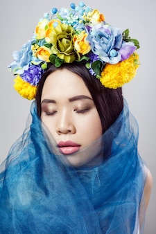 Portrait de jolie femme asiatique avec chapeau floral et voile bleu posant avec les yeux fermés sur fond gris clair. tourné en studio intérieur.