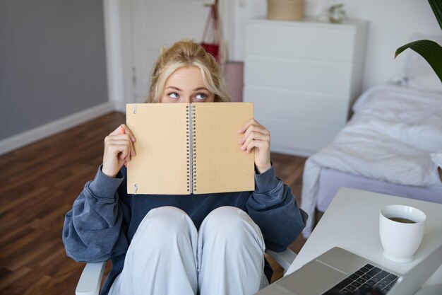 Portrait d'une jolie femme blonde assise devant une caméra numérique et un ordinateur portable dans sa chambre couvre le visage avec