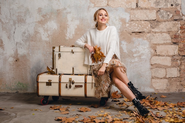 Portrait de jolie femme blonde élégante souriante en pull tricoté blanc assis sur des valises dans la rue contre le mur vintage