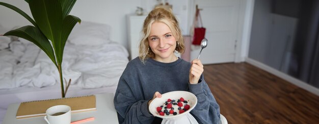 Portrait d'une jolie femme blonde souriante qui prend son petit déjeuner dans sa chambre en regardant un bol avec une caméra.