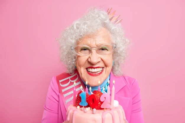 Portrait d'une jolie femme avec du maquillage célèbre son 102e anniversaire souffle des bougies sur un gâteau d'anniversaire sourit avec plaisir porte des vêtements de fête a la fête