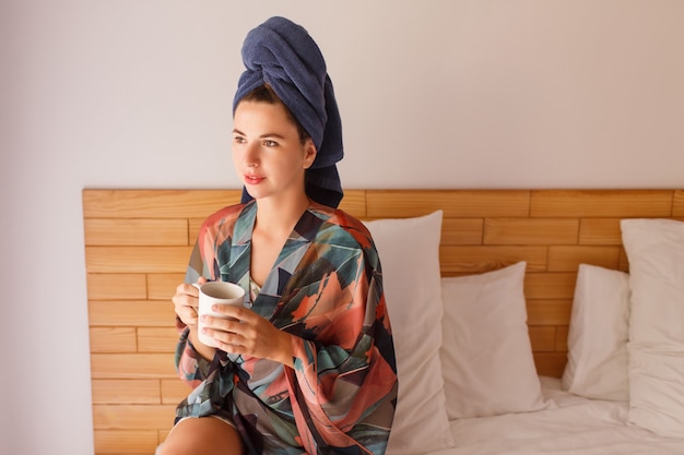 Portrait de jolie femme enveloppée dans une serviette et un peignoir se réveiller le matin assis sur le lit et boire du thé