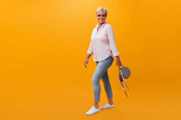 Portrait de jolie femme en jeans et chemise posant avec sac sur fond orange