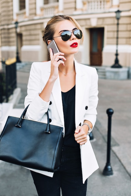 Photo gratuite portrait jolie femme à lunettes de soleil avec des lèvres rouges marchant dans la rue. elle parle au téléphone.