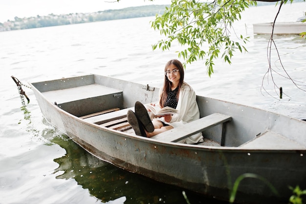 Portrait d'une jolie femme portant une robe à pois noirs, un châle blanc et des lunettes lisant un livre dans un bateau sur un lac