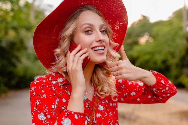 Portrait de jolie femme souriante blonde élégante en chapeau rouge de paille et blouse tenue de mode d'été parler au téléphone geste positif émotion