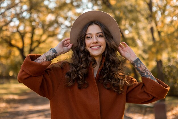 Portrait de jolie femme souriante élégante avec de longs cheveux bouclés marchant dans le parc habillé en manteau brun chaud automne mode tendance, street style wearing hat