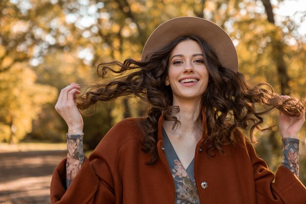 Portrait de jolie femme souriante élégante avec de longs cheveux bouclés marchant dans le parc habillé en manteau brun chaud automne mode tendance, street style wearing hat