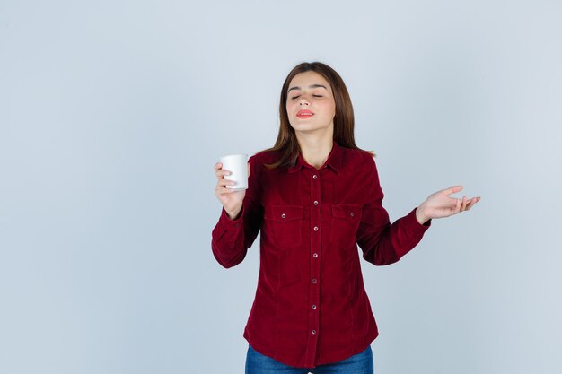 Portrait de jolie femme tenant une tasse de café en plastique tout en fermant les yeux en blouse bordeaux et l'air satisfait