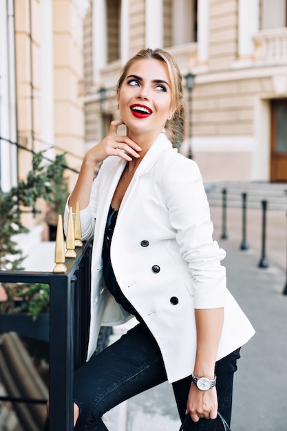 Portrait jolie femme en veste blanche s'appuyant sur une clôture sur la rue. Elle sourit à côté.