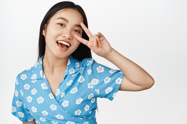 Portrait d'une jolie fille asiatique montre un geste de vsign de paix près des yeux en souriant et en riant avec bonheur à la caméra debout en robe sur fond blanc