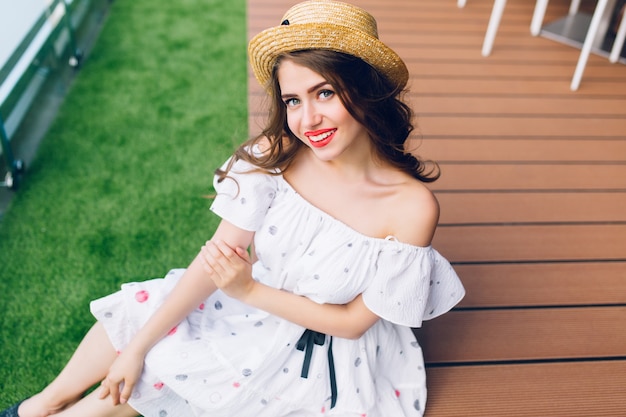 Portrait De Jolie Fille Aux Cheveux Longs Au Chapeau Assis Sur Le Plancher De Bois En Plein Air. Elle Porte Une Robe Blanche Aux épaules Nues, Rouge à Lèvres Rouge. Vue D'en-haut.